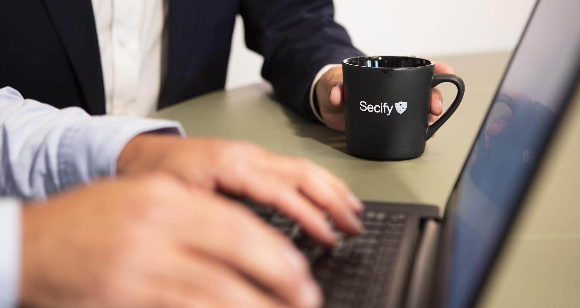 Bärbar dator med kaffekopp i bakgrunden, på koppen står det Secify