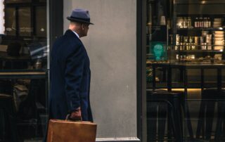 En man går utanför en butik. Mannen har hatt och håller i en brun väska, bär mörka kläder