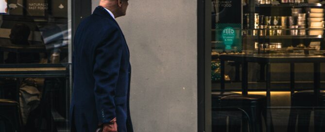 En man går utanför en butik. Mannen har hatt och håller i en brun väska, bär mörka kläder